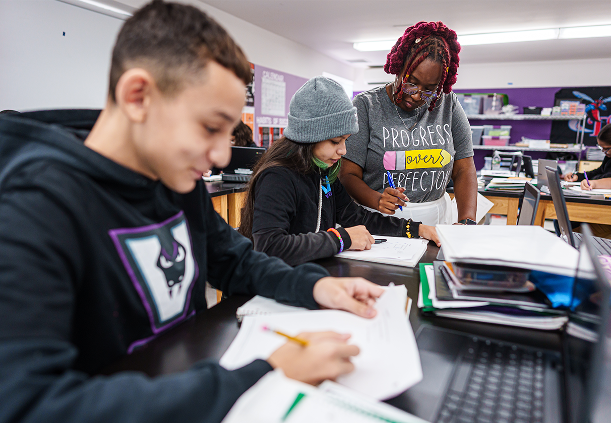 students sitting at desks in a classroom with a teacher standing next to them